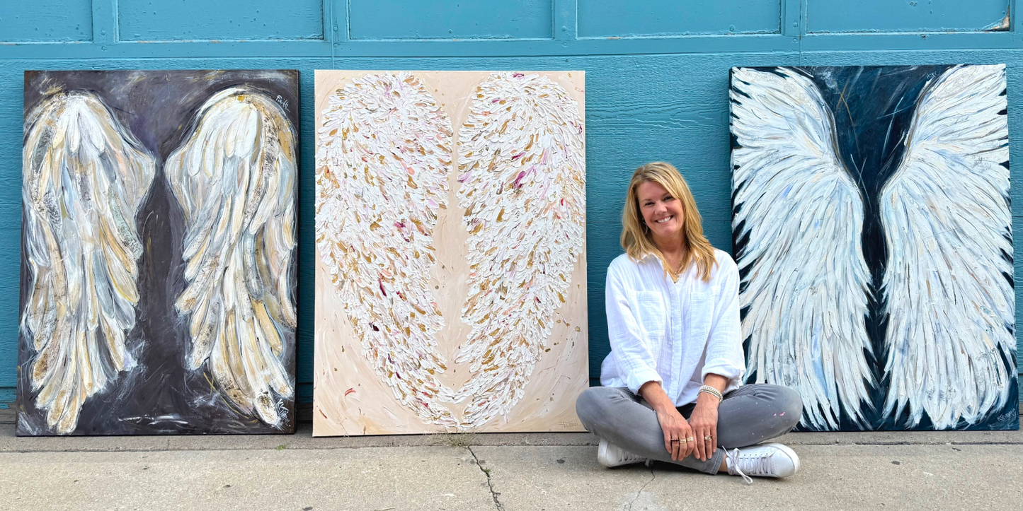 Woman sitting between two large paintings of angel wings against a blue wall.