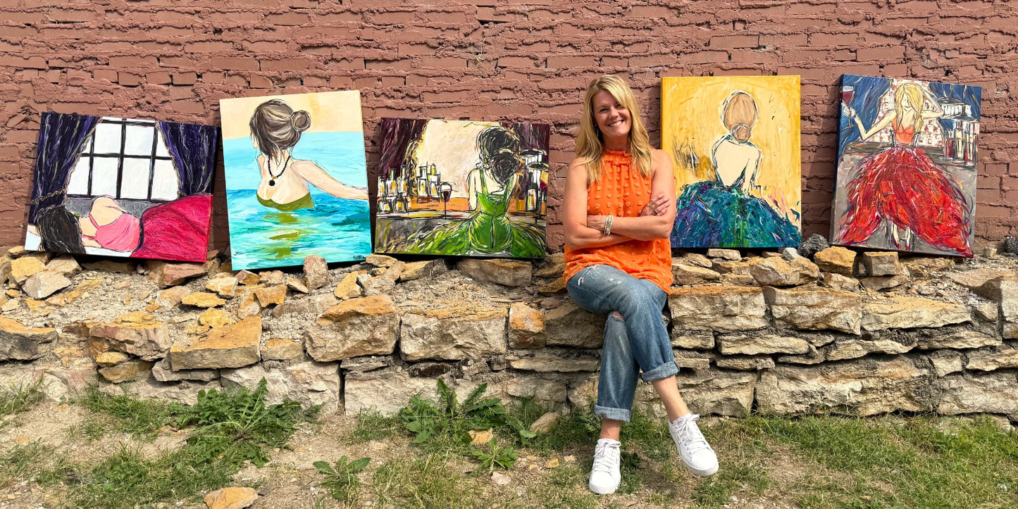 Woman sitting on a stone wall with six colorful paintings behind her against a brick wall.